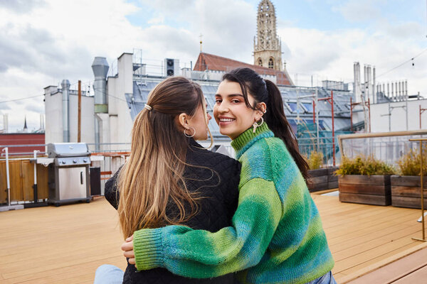 Tender moment between happy lesbian women sitting together on a rooftop, cityscape backdrop