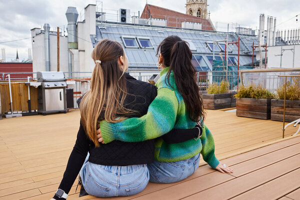 Tender moment between lesbian couple sitting together on a rooftop, cityscape backdrop