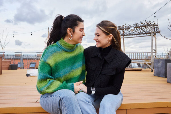 Tender moment between positive lesbian women sitting together on a rooftop, cityscape backdrop