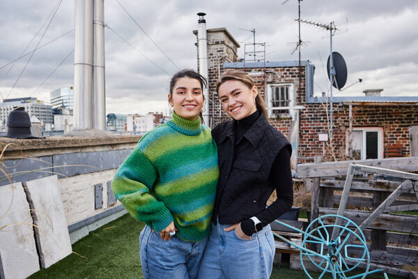 happy young lesbian couple in casual attire standing together of rooftop and looking at camera