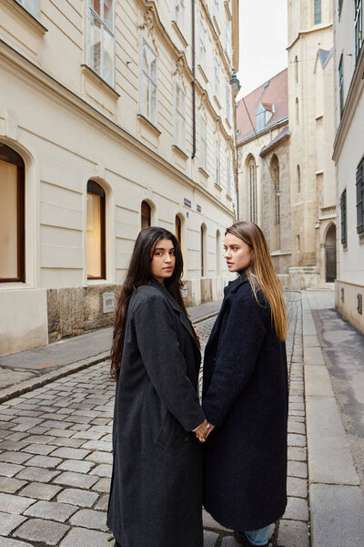 happy young lesbian couple in trendy outerwear holding hands while walking in European city, lgbt