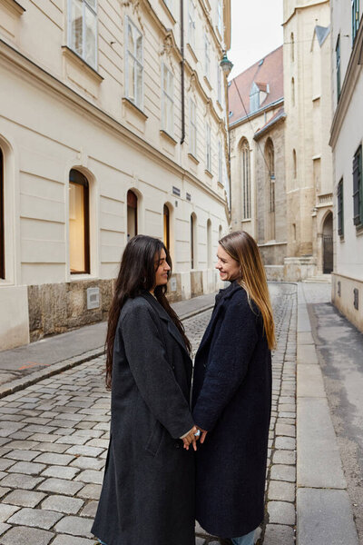 happy young lesbian couple in trendy outerwear holding hands during walk in European city, lgbt
