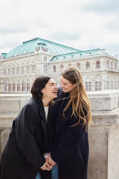 stock image happy lesbian couple in outerwear laughing while standing near building during trip in Vienna