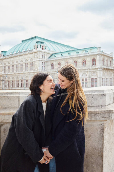 happy lesbian couple in outerwear laughing while standing near building during trip in Vienna