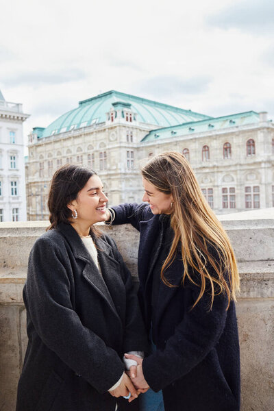 happy lesbian couple in outerwear smiling while standing near building during trip in Vienna