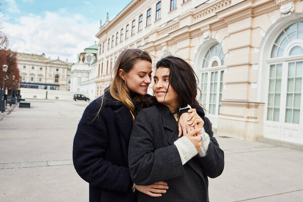 весёлая пара в верхней одежде улыбается, обнимая друг друга возле здания в Вене