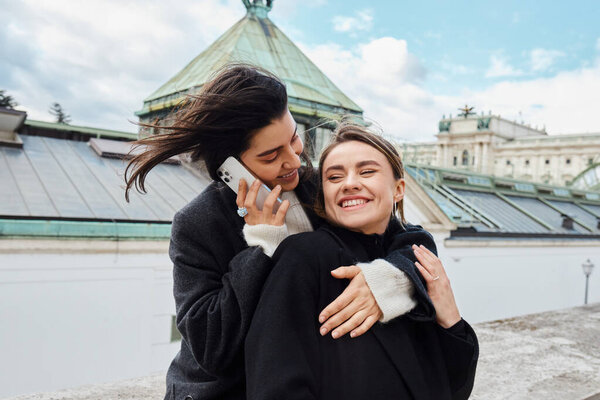 счастливая лесбиянка разговаривает по смартфону и обнимает свою девушку во время поездки в Вену