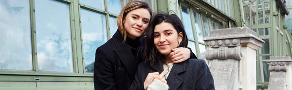 счастливая лесбийская пара в пальто, обнимающая друг друга возле Пальменхауса в Вене на заднем плане, баннер