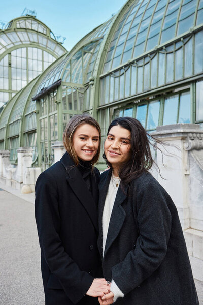 веселая пара в пальто, держась за руки, стоя возле Пальменхауса в Вене на заднем плане