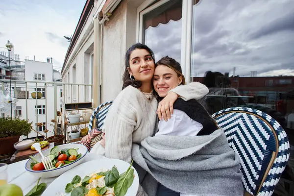 年轻的女同性恋夫妇在阳台上吃早餐时拥抱在一起的快乐时刻 免版税图库照片