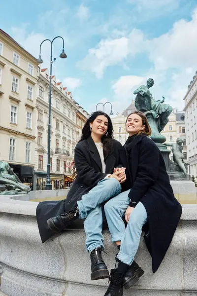 在维也纳 一对快乐的女同性恋夫妇手牵着手 坐在一座有着城市景观的喷泉雕像旁边 图库图片