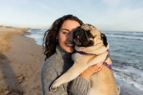 Cheerful and curly young woman holding pug dog on beach near sea in Barcelona — Stock Photo
