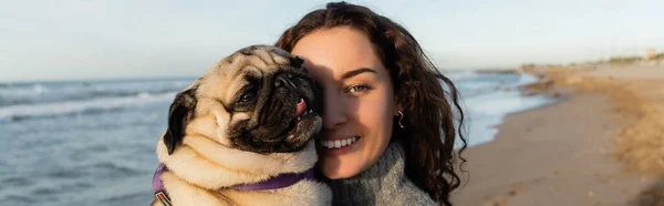 Cheerful and curly young woman holding pug dog on beach near sea, banner — Stockfoto
