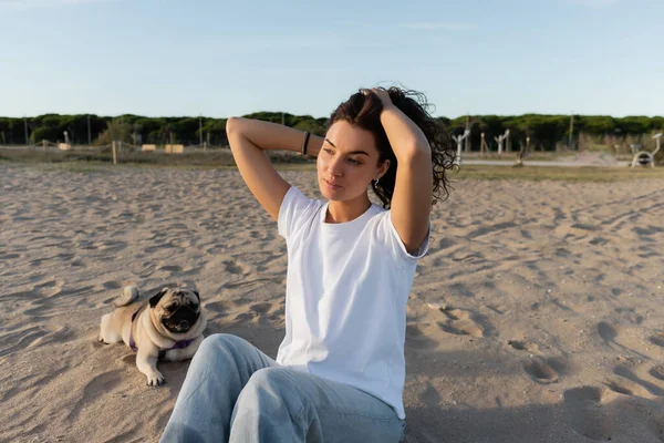 Young woman adjusting curly hair while sitting near pug dog on sandy beach in Barcelona — Photo de stock