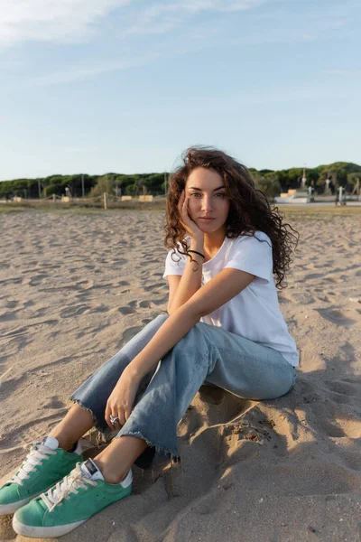 Jeune femme bouclée assise en jeans et t-shirt blanc sur une plage de sable fin à Barcelone — Photo de stock