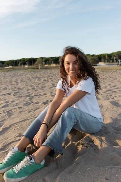 Cheerful young woman sitting in jeans and white t-shirt on sandy beach in Barcelona — Stock Photo