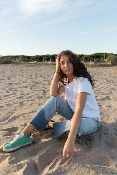 Curly woman sitting in blue jeans and white t-shirt on sandy beach in Barcelona — Stock Photo