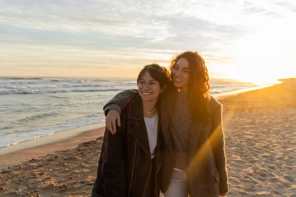 Cheerful woman hugging friend and smiling while walking during sunset on sandy beach — Stock Photo