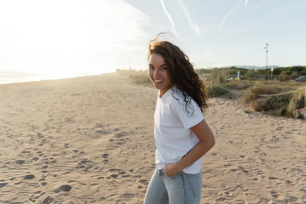 Cheerful young woman in white t-shirt walking on sandy beach in Spain — стокове фото