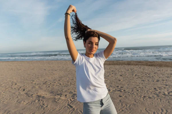 Brunette woman in white t-shirt adjusting curly hair on sandy beach near sea — Stock Photo