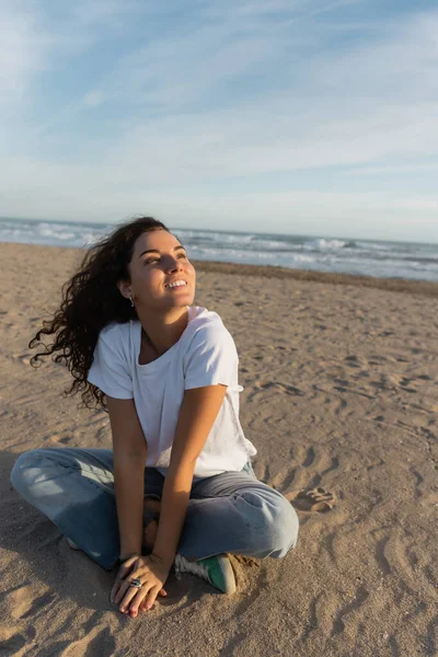 Dreamy woman sitting in blue jeans and white t-shirt sitting on sandy beach in Spain — Stock Photo