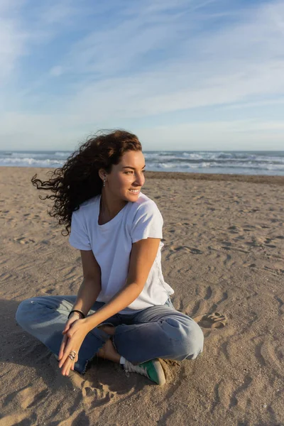 Positive woman sitting in blue jeans and white t-shirt sitting on sandy beach in Spain — Photo de stock