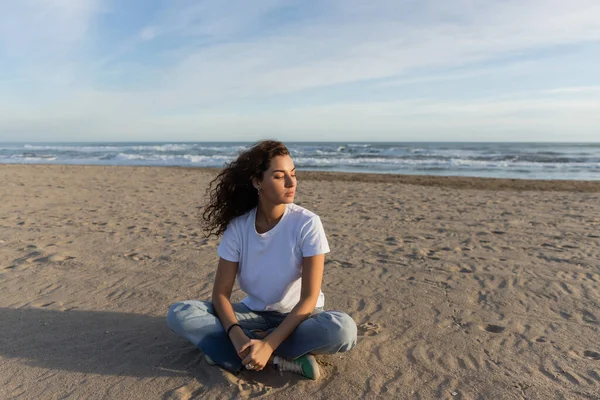 Кудрявая брюнетка, сидящая в синих джинсах и белой футболке на песчаном пляже Испании — стоковое фото