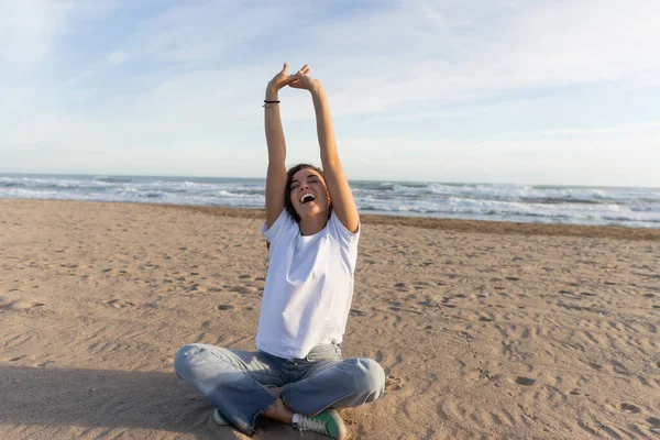 Mujer positiva sentada en jeans azules y camiseta blanca sentada con las manos levantadas en la playa de arena en España - foto de stock