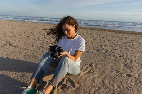 Кудрявая женщина сидит в синих джинсах и белой футболке, сидя с цифровой камерой на пляже в Испании — стоковое фото