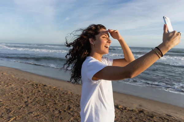 Femme bouclée en t-shirt blanc prenant selfie sur smartphone sur une plage de sable fin à Barcelone — Photo de stock
