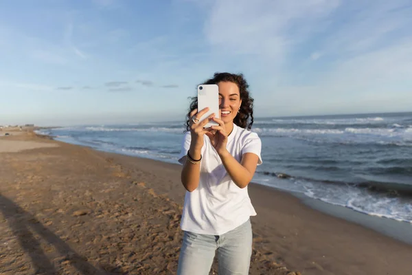 Cheerful woman in white t-shirt taking photo on smartphone on sandy beach in Barcelona — Stock Photo