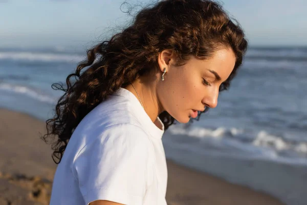 Portrait of brunette curly woman in white t-shirt on beach near sea — Photo de stock