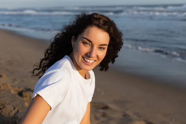 Portrait of positive and curly woman in white t-shirt on sandy beach near sea — Stock Photo