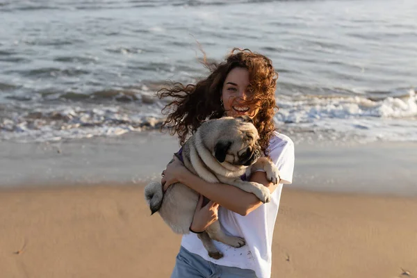 Fröhliche junge Frau mit lockigem Haar hält Mops am Meer in Spanien — Stockfoto