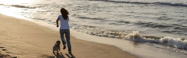 Vue arrière de la femme courant avec chiot chiot sur la plage de sable fin pendant le coucher du soleil, bannière — Photo de stock