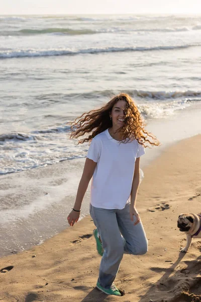 Весела молода жінка біжить з мопсом на піщаному пляжі під час заходу сонця — стокове фото
