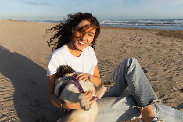Cheerful young woman with curly hair holding pug dog while sitting on sand near sea in Spain — Stock Photo