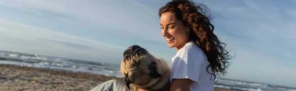 Feliz joven con pelo rizado sosteniendo perro pug mientras está sentado en la playa cerca del mar en España, pancarta - foto de stock
