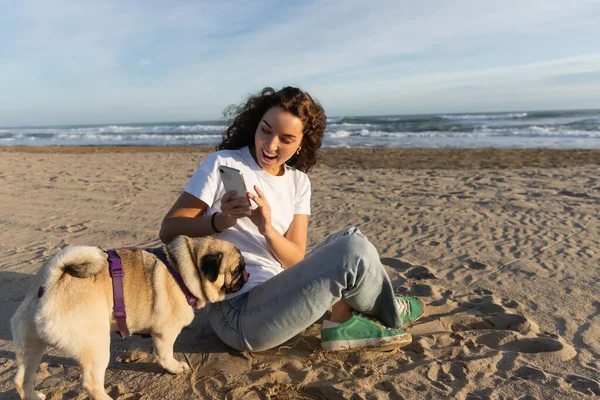 Cheerful woman in white t-shirt taking photo of pug dog on sandy beach in Barcelona — Stockfoto
