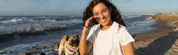 Cheerful young woman covering cheek from sunshine near pug dog on beach near sea in Spain, banner — Foto stock