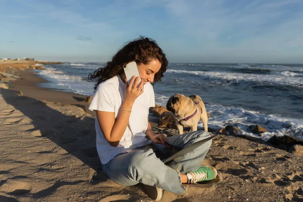 Alegre freelancer hablando en smartphone mientras usa el ordenador portátil cerca de perro pug en la playa cerca del mar en España - foto de stock