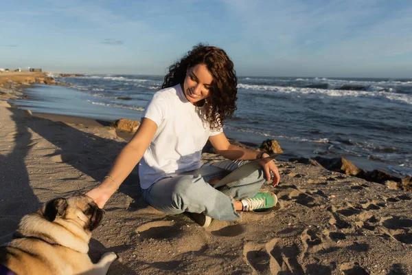 Cheerful freelancer sitting with laptop and cuddling pug dog on beach near sea in Spain — Stockfoto