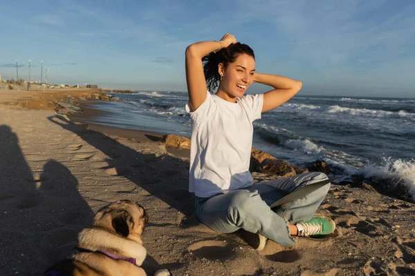 Fröhliche Freiberuflerin sitzt mit Laptop und lockigem Haar in der Nähe von Mops Hund am Strand am Meer in Spanien — Stockfoto