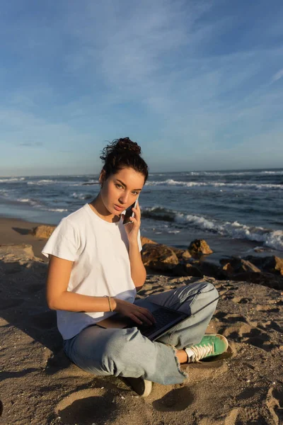 Young freelancer sitting with laptop and talking on smartphone near sea in Spain — Stock Photo