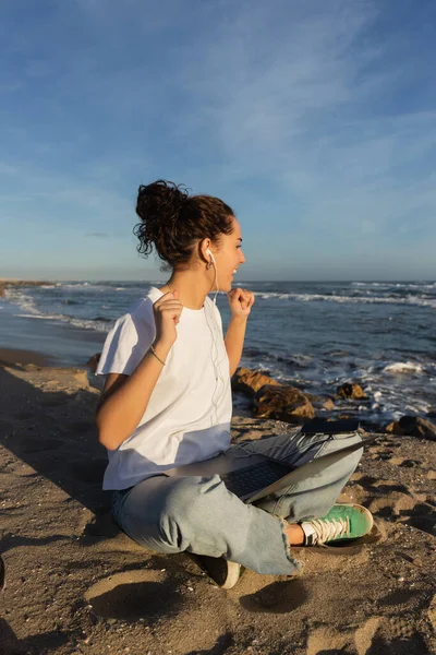 Feliz jovem freelancer em fones de ouvido com fio sentado com laptop na praia em Barcelona — Fotografia de Stock