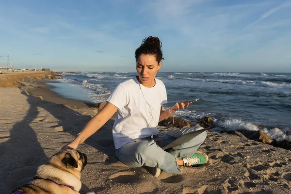 Young woman in wired earphones holding smartphone near laptop and cuddling pug dog on beach in Barcelona — Photo de stock