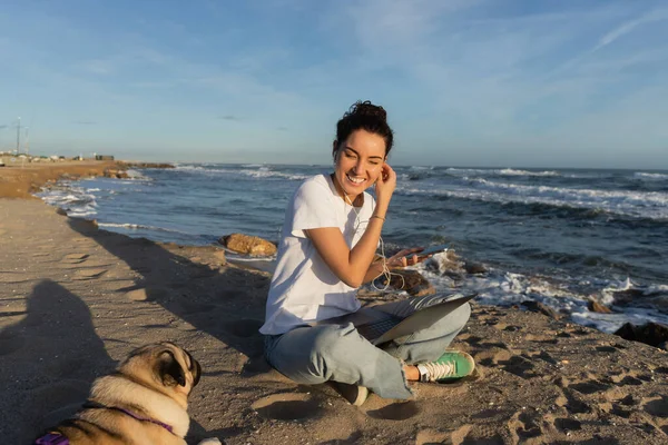 Happy woman wearing wired earphones while holding smartphone near laptop and pug dog on beach in Barcelona — Stock Photo