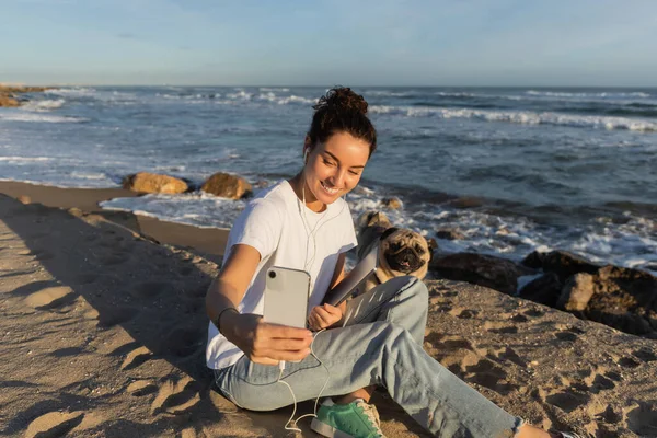 Cheerful woman in wired earphones holding laptop and taking selfie with pug dog near sea in Spain — Stock Photo