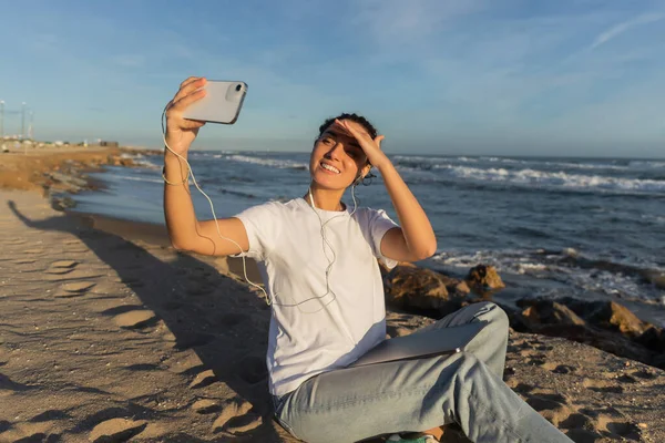 Cheerful woman in wired earphones sitting with laptop and taking selfie near sea in Spain — Fotografia de Stock