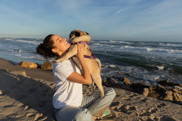 Positive junge Frau mit geschlossenen Augen hält Mops Hund am Strand in der Nähe des Meeres in Barcelona — Stockfoto